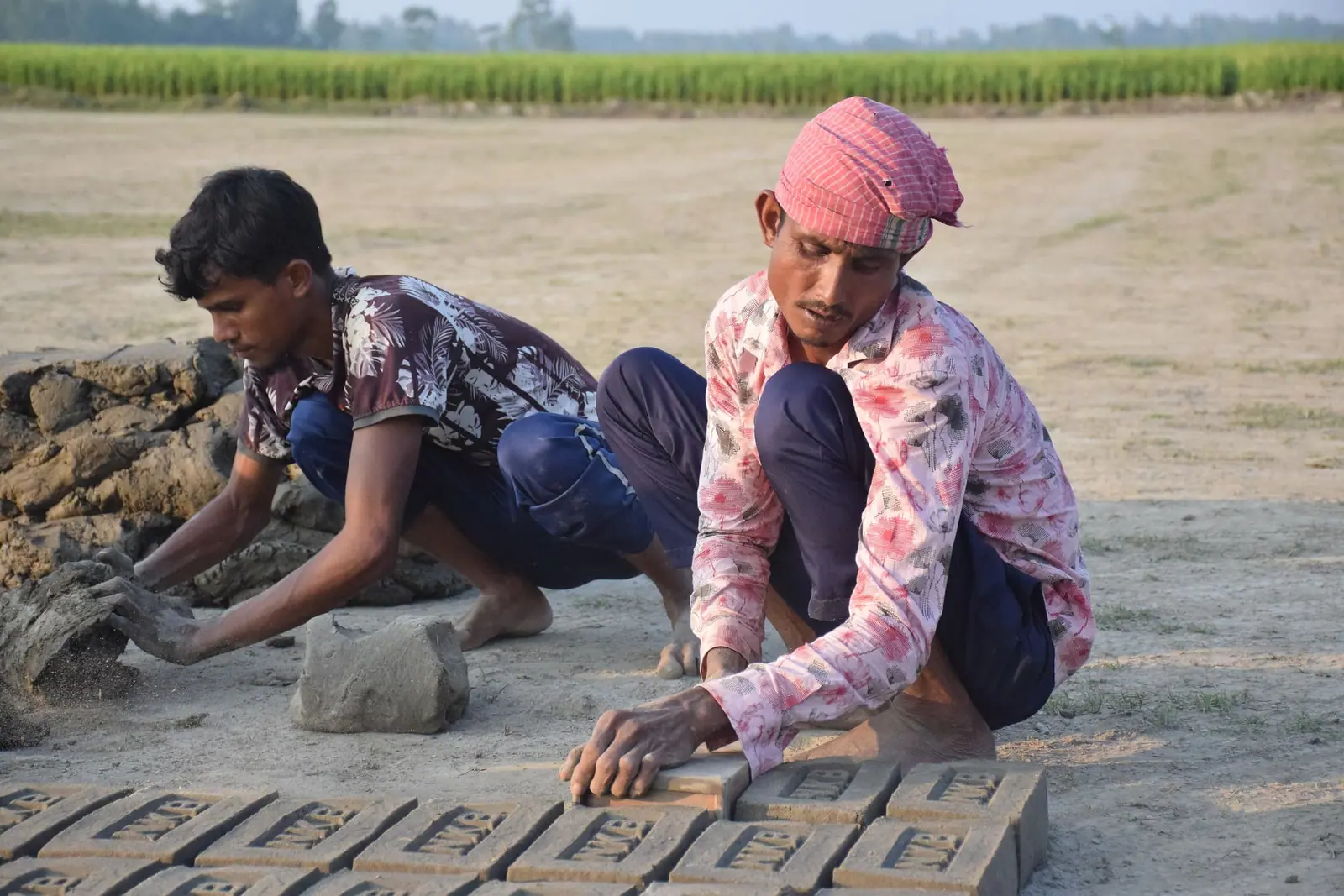 Two brick makers kneeling on the ground, pressing wet clay into moulds beside a row of shaped bricks