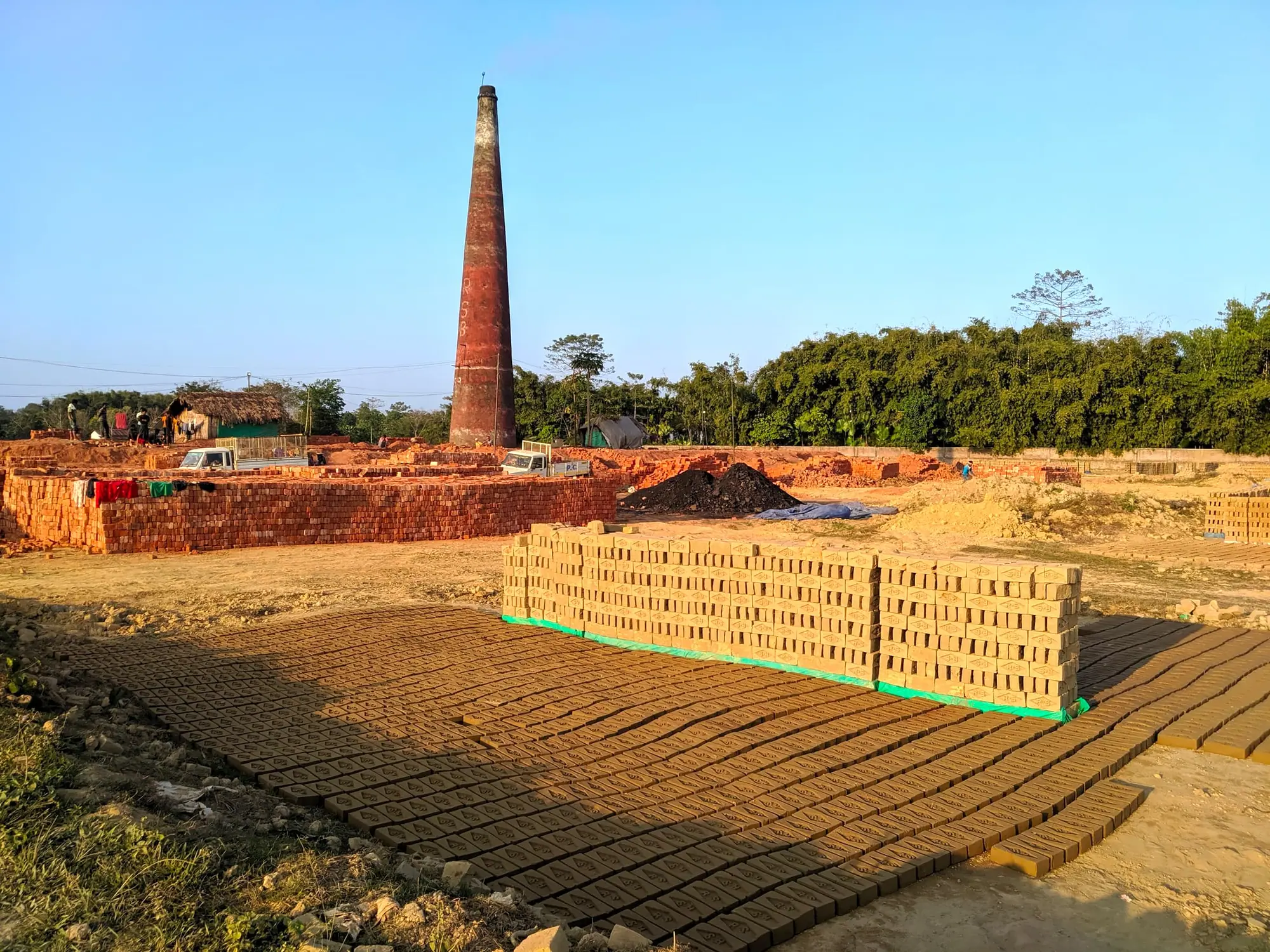 Rows of freshly moulded clay bricks laid out to dry in front of a tall brick-kiln chimney at golden hour