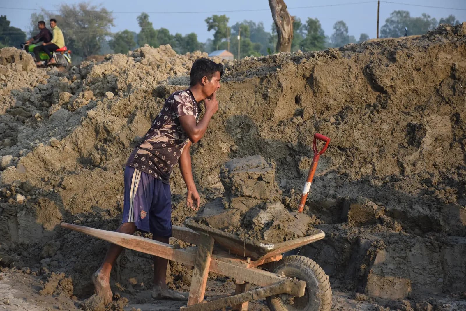 A worker pushing a wheelbarrow of raw clay past a freshly dug clay bank
