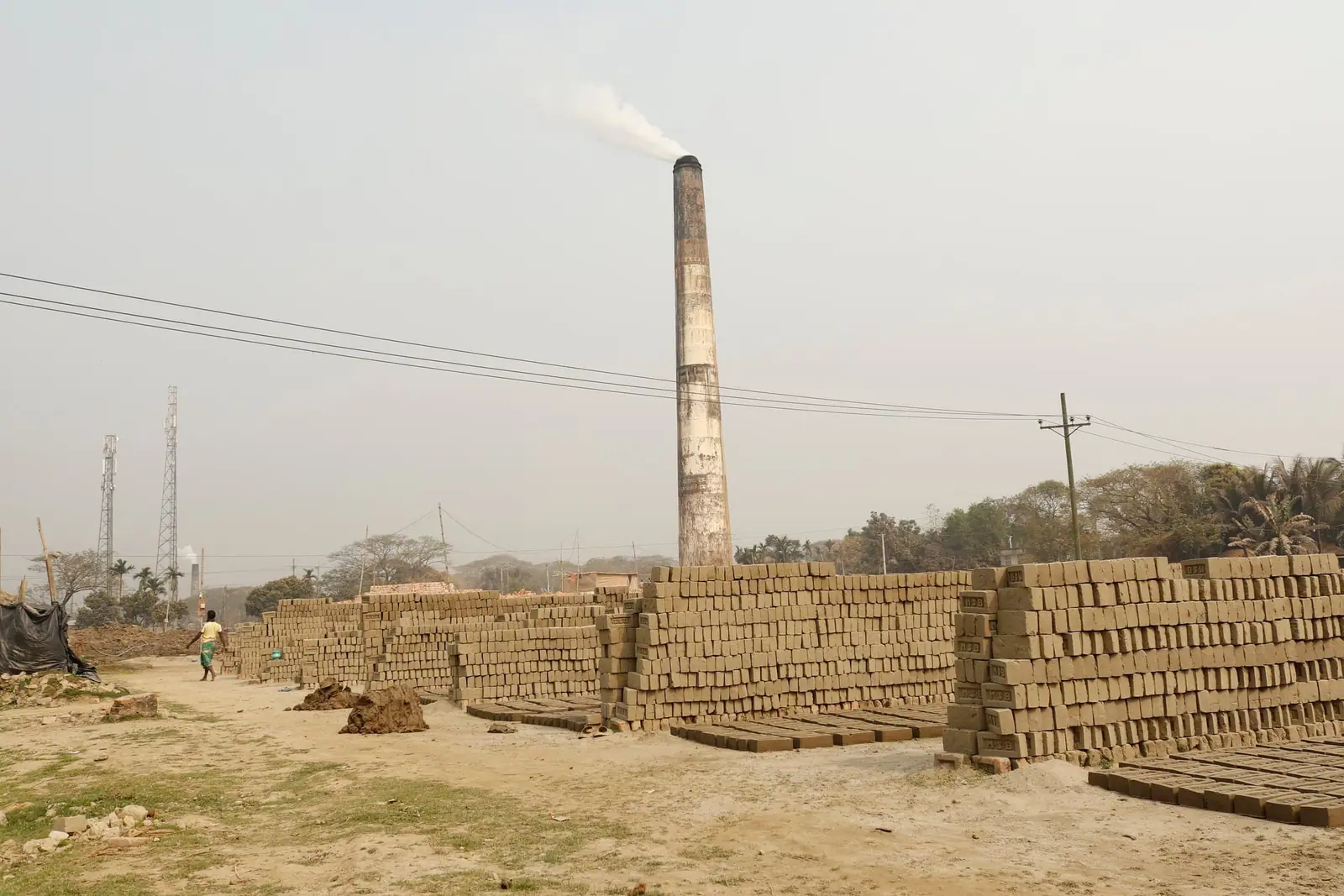 Stacked bricks in a brick field with a tall kiln chimney releasing smoke in the background