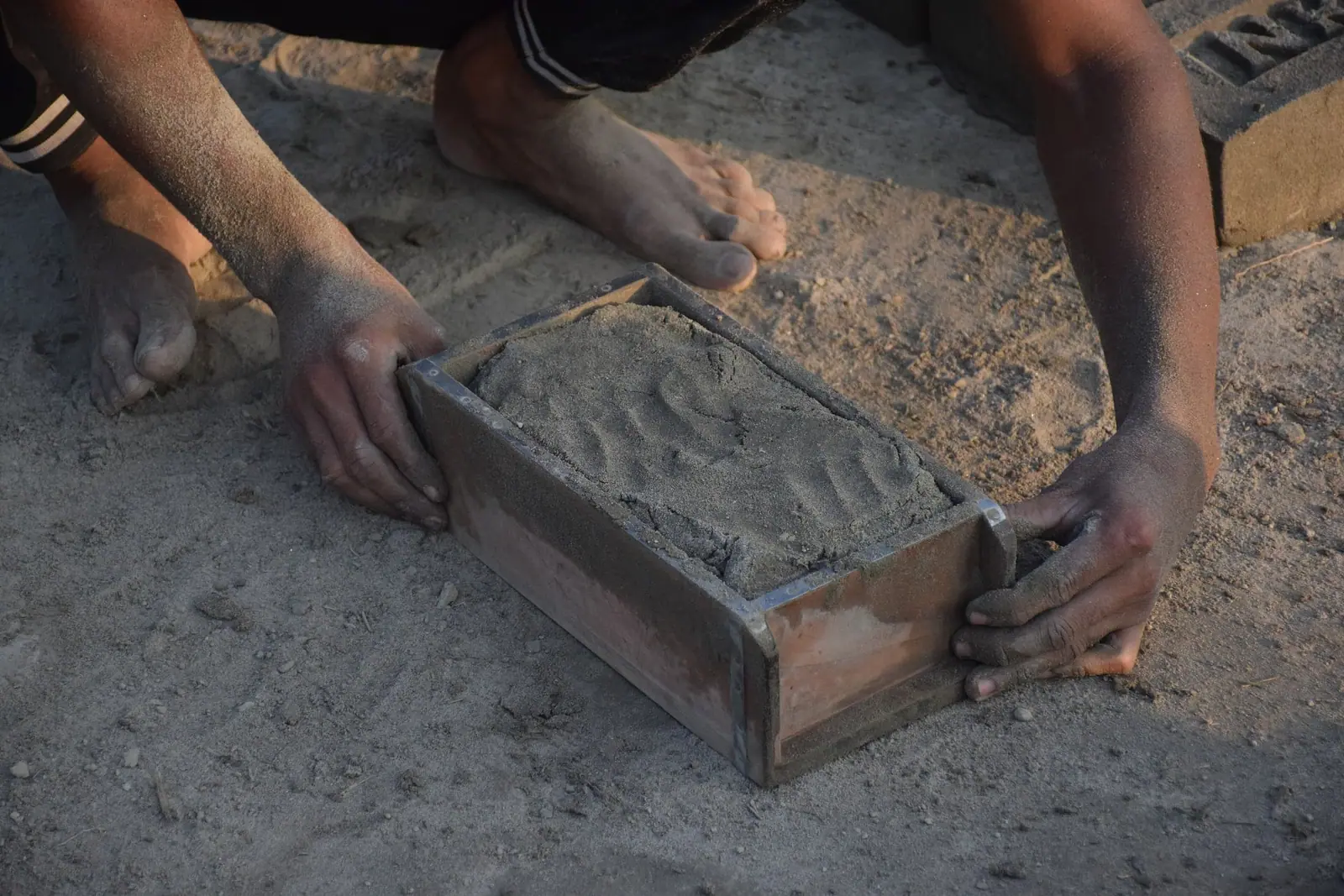 Hands pressing wet clay into a wooden brick mould on the ground